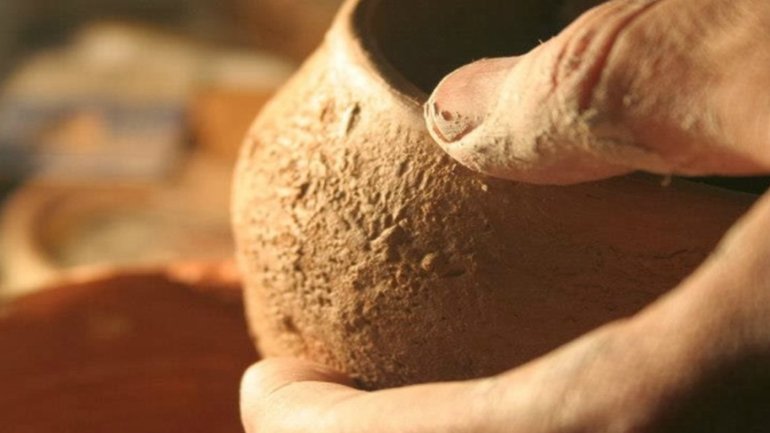 Primer pla d'unes mans sostenint una peça de ceràmica de fang en procés de modelatge, en un taller amb eines desenfocades al fons.