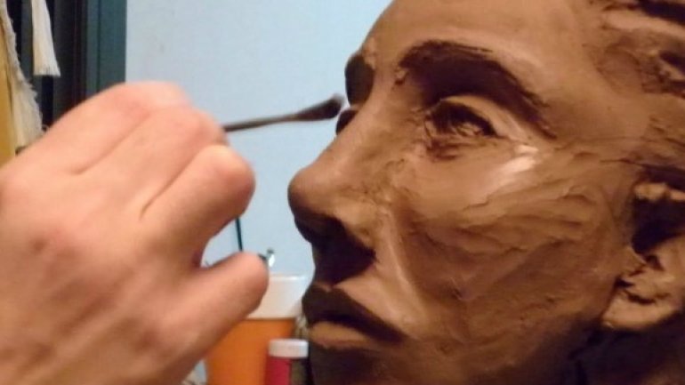 Una mà modela amb una eina un bust d'argila de grans dimensions en un taller d'escultura. La imatge mostra el procés creatiu i el treball de detall sobre el rostre.