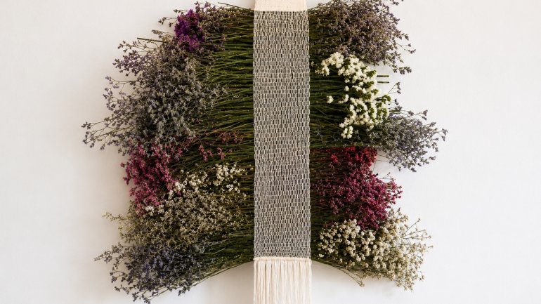 Tapís tèxtil amb flors seques disposades en capes i una franja central blanca amb serrells, penjat sobre una paret clara.