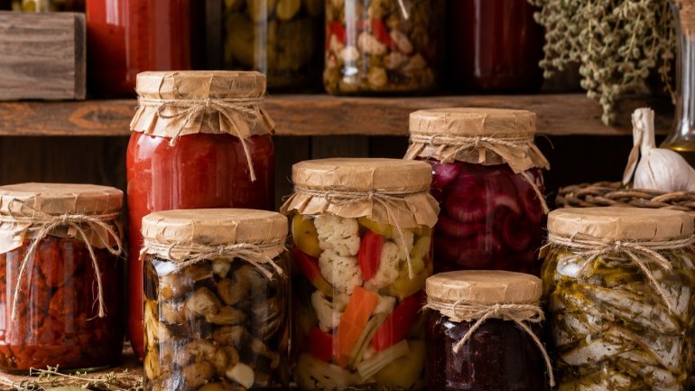 En aquesta imatge es veuen diferents pots de vidre plens d'hortalises en conserva en un ambient rústic. 
