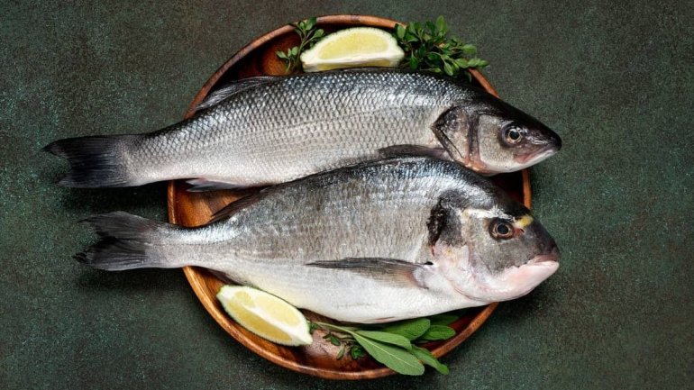 A la imatge es veuen dos peixos sencers col·locats sobre una safata rodona de fusta, amb una presentació típica de peix fresc.
El peix de dalt sembla un llobarro: té el cos allargat, de color gris platejat, amb escates visibles i una cua lleugerament fos