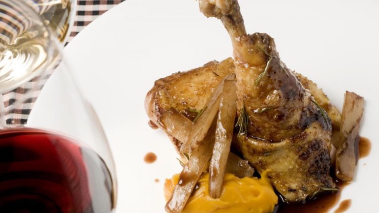 Cuixa de gall rostida servida amb puré de carbassa, ceba rostida i salsa, presentada en un plat blanc amb una copa de vi negre i una de vi blanc al costat.
