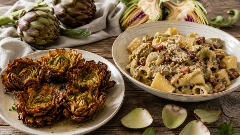 Plat de pasta amb carxofes, cansalada i formatge ratllat, acompanyat de carxofes rostides i carxofes fresques sobre una taula de fusta.