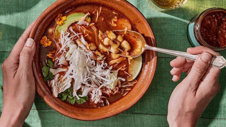 Bol de pozole mexicà amb brou vermell, blat de moro cacauazintle, col blanca, alvocat, coriandre i llima, sostingut amb les mans.