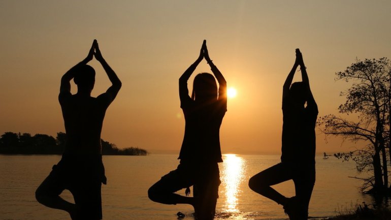 Tres persones practiquen ioga a la vora d'un llac durant la posta de sol. Es mantenen dretes sobre una cama, amb l'altra flexionada i les mans unides per sobre del cap en una postura d'equilibri. Les seves siluetes es retallen contra el cel daurat i el re