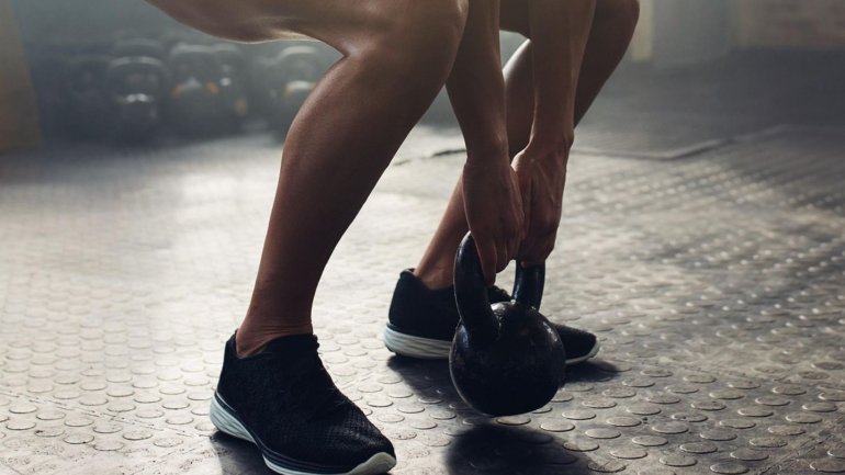 La imatge mostra una persona en un gimnàs agafant una pesa russa (kettlebell) mentre flexiona les cames per iniciar un exercici. Es destaca la força i la definició muscular de les cames, així com un ambient esportiu que transmet esforç, concentració i act