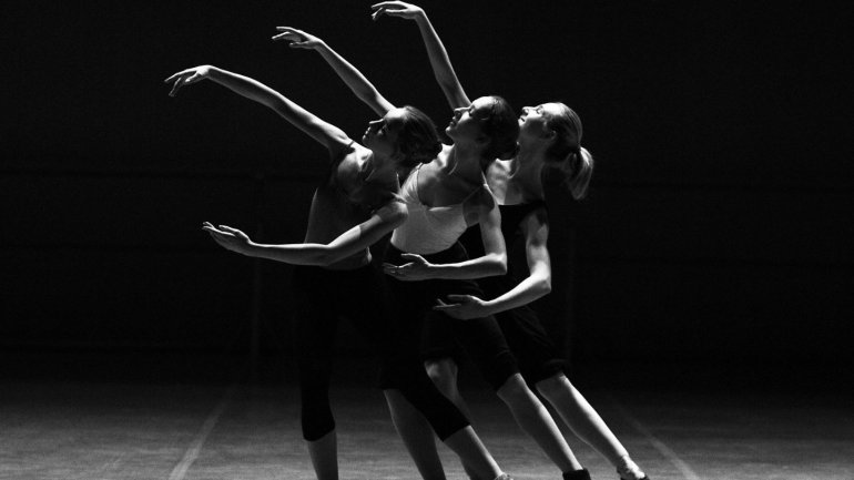 Tres ballarines interpreten una coreografia sobre un escenari fosc. Amb els cossos inclinats i els braços estesos cap amunt i als costats, formen una composició harmoniosa que transmet moviment i equilibri.