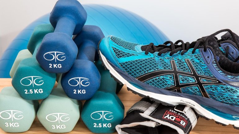 Sobre una superfície de fusta hi ha diversos elements per fer exercici físic. A l'esquerra, unes manuelles de diferents pesos, de color blau i verd, estan apilades davant d'una pilota d'exercici blava. A la dreta, unes sabatilles esportives de color blau 