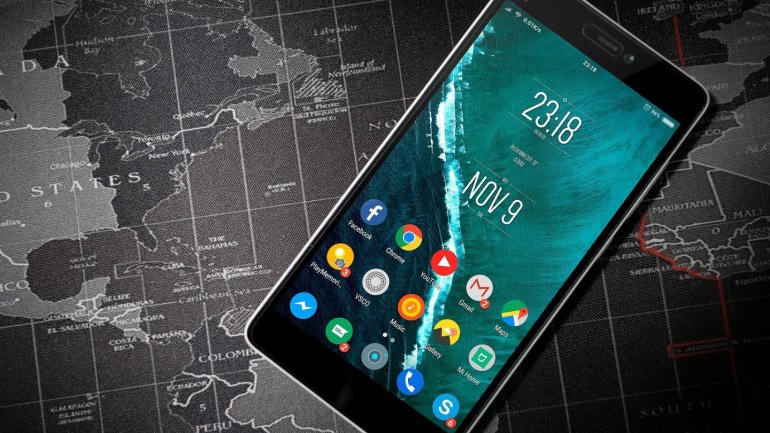 Un telèfon intel·ligent amb la pantalla encesa descansa sobre un mapa del món en tons grisos. A la pantalla s'hi veuen diverses aplicacions, l'hora (23:18) i la data (9 de novembre). La imatge relaciona la tecnologia i la comunicació amb la connexió globa