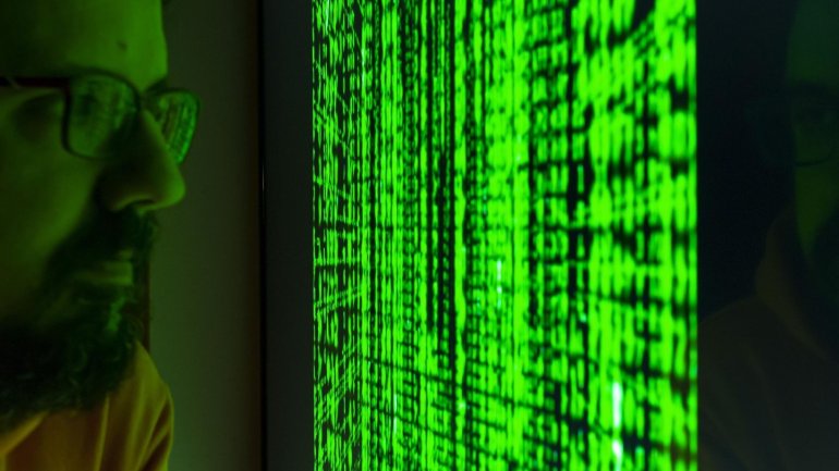 Una persona amb ulleres observa una pantalla que mostra una gran quantitat de caràcters verds sobre un fons fosc. La pantalla il·lumina parcialment el seu rostre, mentre la resta de l'espai queda en penombra.