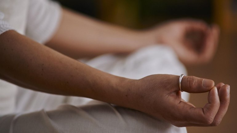 Una persona està asseguda en postura de meditació, amb les mans reposant sobre els genolls i els dits formant un gest de relaxació. La imatge se centra en les mans i els braços, mentre la resta del cos apareix desenfocada. Vesteix roba clara i l'ambient é