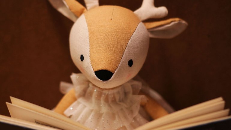 Un peluix amb forma de cérvol està situat darrere d'un llibre obert, com si l'estigués llegint. Té el cos de roba en tons beix i blanc, petites banyes, orelles grans i un volant de tul al coll. El fons és llis i de color marró, cosa que fa destacar la fig