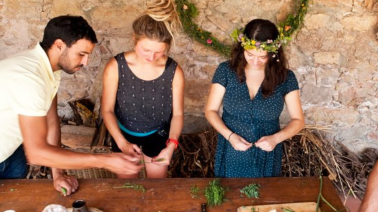 Tres persones al voltant d'una taula realitzant una activitat, manipulant plantes silvestres, en un espai rústic.