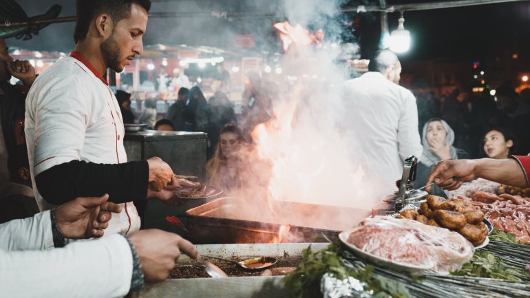 Parada de menjar de carrer en un mercat nocturn, amb diversos cuiners preparant carn a la graella sobre flames vives i fum. En primer pla hi ha broquetes, salsitxes, herbes i altres ingredients, mentre clients observen i esperen el seu menjar al fons.
