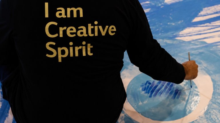 Persona participant en una activitat artística pintant sobre un llenç amb tons blaus i blancs, mostrant una samarreta negra amb el missatge “I am Creative Spirit” en un espai creatiu.