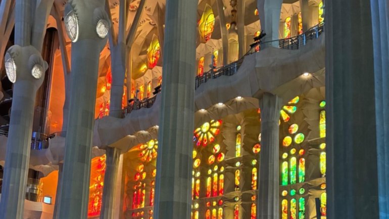 Interior de la Basílica de la Sagrada Família, amb les altes columnes inspirades en arbres i els vitralls de colors que projecten una llum intensa en tons vermells, taronges, grocs i verds, creant una atmosfera lluminosa i monumental.