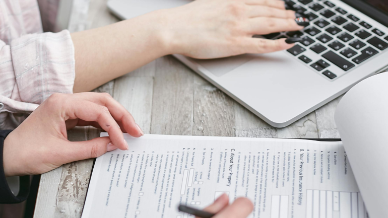 imatge de  dos persones on només es veuen les mans que estan omplin un formulari referent a un contracte editorial. sobre un escriptori amb un ordinador i pepers.