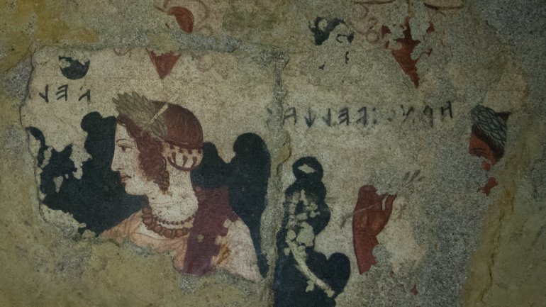 Fragment d’un fresc antic sobre una paret envellida, amb la representació d’una dona de perfil amb pentinat i joies d’estil clàssic, envoltada de restes de figures i inscripcions d’època antiga.
