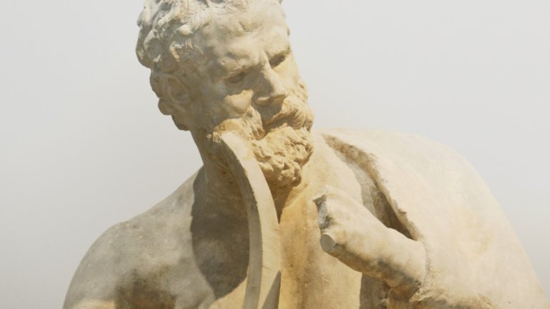 Primer pla d'una escultura de marbre que representa Homer, el llegendari poeta de l'antiga Grècia, amb barba i expressió reflexiva, il·luminada per la llum natural sobre un fons clar.