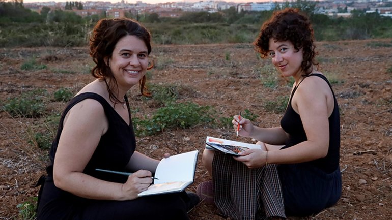 Les professores Mònica Kopatschek i Ana López Serrano, assegudes a terra en un camp, pinten amb aquarel·la durant una sessió a l'aire lliure. Totes dues miren cap a la càmera i somriuen. Al fons es veu un paisatge urbà sota la llum del capvespre.
