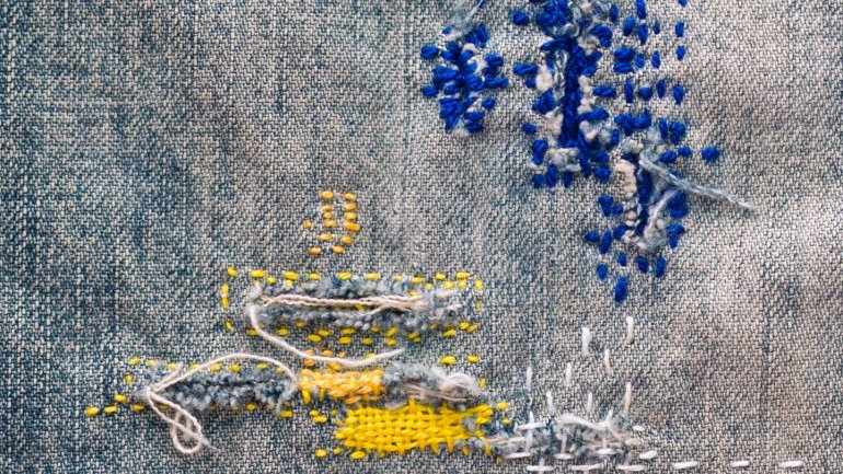Brodat contemporani sobre lli gris amb tècniques de visible mending, combinant punts decoratius, reparacions tèxtils i textures en fils blaus, grocs i blancs.