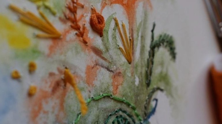 Detall d’un brodat contemporani sobre tela, amb fils de colors que ressegueixen un motiu vegetal pintat amb aquarel·la.