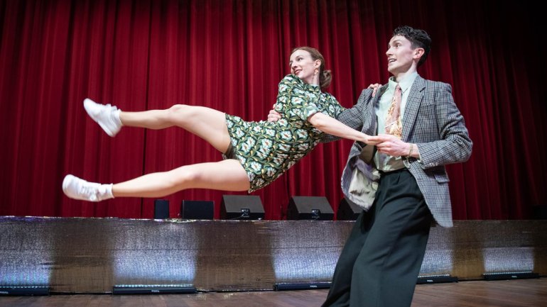 Parella realitzant una figura acrobàtica de dansa sobre un escenari amb un teló vermell de fons