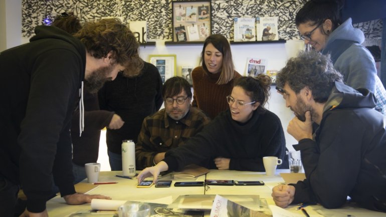 Grup de participants reunits al voltant d'una taula en un taller de fotografia i cartografia sensible, observant imatges i materials de treball mentre comparteixen idees i analitzen el procés creatiu en un ambient col·laboratiu.