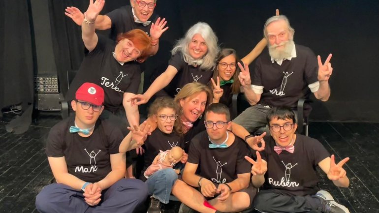 Grup de participants d’un projecte de teatre inclusiu posant junts sobre l’escenari, amb expressions alegres i gestos divertits, davant d’un teló negre.