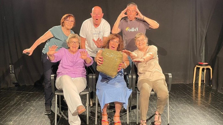 Grup de sis persones participant en una activitat de teatre, representant una escena amb gestos expressius i divertits sobre un escenari amb teló negre. Una de les participants sosté un objecte mentre la resta interactua amb ella.