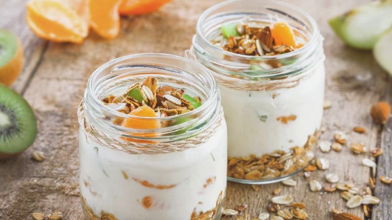 Dos pots de vidre amb iogurt, capes de granola i trossos de fruita, sobre una taula de fusta rústica. Al voltant hi ha kiwi, taronja, fruits secs i cereals.
